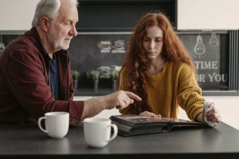 An older man and his adult daughter look through a photo album at a kitchen table over coffee