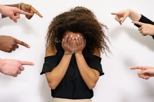 A woman hides her face in her hands while several hands point at her from both sides.