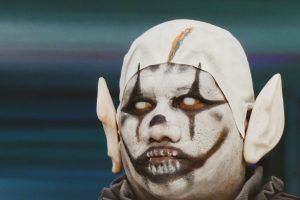 A person in white clown makeup with pointed prosthetic ears and a grey ruffled collar.